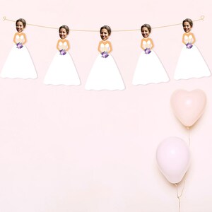 May include: A bridal shower banner featuring five white dresses with purple flowers and a bride's face in each dress. The banner is strung with twine and there are two pink balloons in the background.