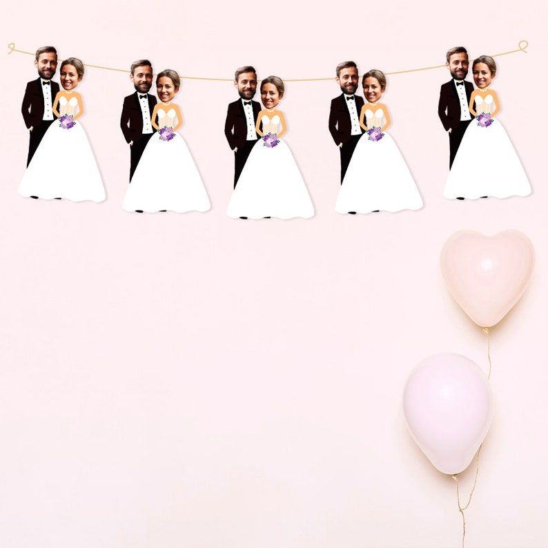 May include: A wedding banner with a string of bride and groom cutouts. Each cutout features a photo of a couple's faces. The banner is strung with white twine and hangs against a light pink background. Two pink balloons are in the bottom right corner of the image.