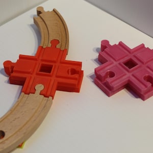 May include: A wooden curved train track section with a red plastic connector and a separate pink plastic track connector. The track is light brown, and the connectors are bright red and pink. Toy train accessories.