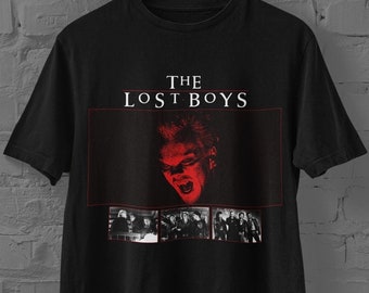 Puede incluir: Una camiseta negra de manga corta con el texto THE LOST BOYS en letras blancas. Debajo del texto hay un gráfico en tonos rojos de un rostro con colmillos y tres imágenes rectangulares en blanco y negro de escenas de una película.