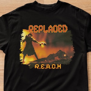 以下が含まれることがあります： 黒いTシャツ。オレンジ色のピクセル化された文字で「REPLACED」と書かれ、その上に武器を持った人物のピクセル化されたグラフィックがあります。「R.E.A.O.H」という文字が下にあります。Tシャツは木の床に置かれています。