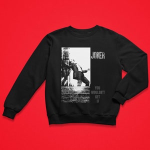 Op de afbeelding: Zwart sweatshirt met een zwart-wit afbeelding van een persoon in een pak, met het woord "JOKER" erboven en "YOU WOULDN'T GET IT" eronder. Het sweatshirt wordt gepresenteerd tegen een rode achtergrond.