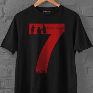 Puede incluir: Una camiseta negra de manga corta con un gráfico grande del número siete en color rojo. El diseño incluye siluetas de tres figuras y el texto "What's in the box?" y "se7en" impresos en rojo. La camiseta tiene cuello redondo y está en una percha.