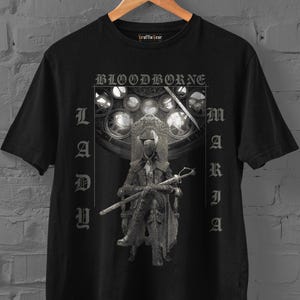May include: A black short-sleeve t-shirt featuring a graphic print of a figure sitting on a throne. The text "BLOODBORNE" appears at the top, with "LADY" and "MARIA" printed vertically on the sides. The design includes a circular window background.