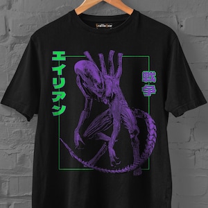 Puede incluir: Una camiseta negra de manga corta con un gráfico morado de una criatura xenomorfa centrado dentro de un marco rectangular verde fino. El diseño incluye texto japonés verde en los lados izquierdo y derecho del marco sobre un fondo oscuro.