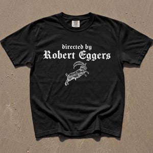 May include: A black short-sleeve t-shirt featuring white text that reads directed by Robert Eggers in a gothic font. Below the text is a detailed white line-art illustration of a leaping goat with curved horns, centered on the chest area.