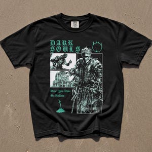 May include: A black short-sleeve t-shirt featuring a teal graphic print. The design includes the text "DARK SOULS" and "Don't You Dare Go Hollow," along with illustrations of a knight in armor, a gothic building, and a sword in the ground.