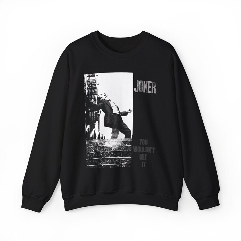 Op de afbeelding: Zwart sweatshirt met een grijswaardenafbeelding van een persoon die tegen een gebouw leunt. Het woord "JOKER" staat erboven en "YOU WOULDN'T GET IT" eronder. Het sweatshirt is gemaakt van zacht materiaal.