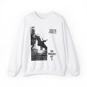Op de afbeelding: Witte sweatshirt met een zwart-wit afbeelding van een figuur die tegen een gebouw leunt, met het woord "JOKER" erboven en "YOU WOULDN'T GET IT" eronder. De sweatshirt heeft een ronde hals en lange mouwen.