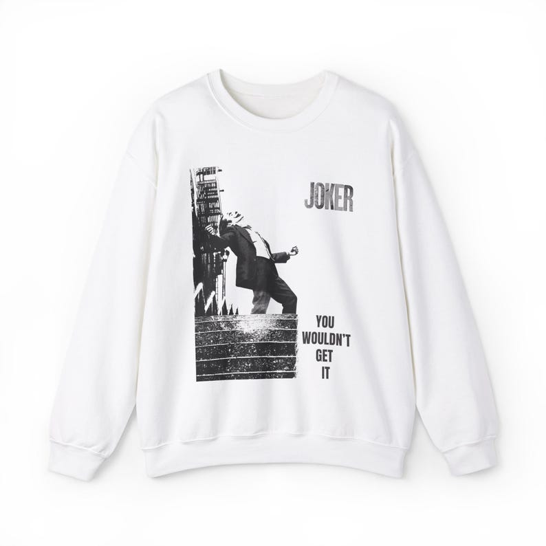 Op de afbeelding: Witte sweatshirt met een zwart-wit afbeelding van een figuur die tegen een gebouw leunt, met het woord "JOKER" erboven en "YOU WOULDN'T GET IT" eronder. De sweatshirt heeft een ronde hals en lange mouwen.