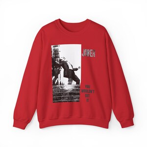 Op de afbeelding: Een rode sweatshirt met een zwart-wit afbeelding van een man die tegen een gebouw leunt. Het woord "JOKER" is in zilver gedrukt en de tekst "YOU WOULDN'T GET IT" staat onder de afbeelding.