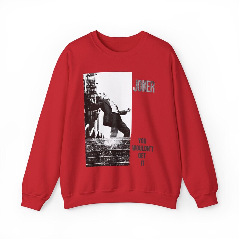Op de afbeelding: Een rode sweatshirt met een zwart-wit afbeelding van een man die tegen een gebouw leunt. Het woord "JOKER" is in zilver gedrukt en de tekst "YOU WOULDN'T GET IT" staat onder de afbeelding.