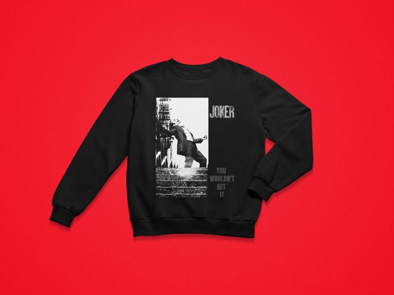 Op de afbeelding: Zwart sweatshirt met een zwart-wit afbeelding van een persoon in een pak, met het woord "JOKER" erboven en "YOU WOULDN'T GET IT" eronder. Het sweatshirt wordt gepresenteerd tegen een rode achtergrond.
