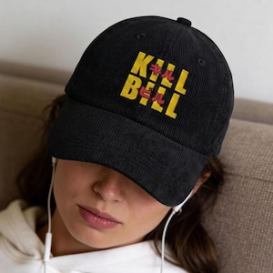May include: A black corduroy baseball cap featuring the yellow text KILL BILL embroidered on the front, with red Japanese katakana characters positioned between the letters. The cap has a structured crown, a curved brim, and a button on top.