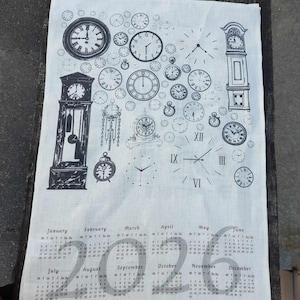 Könnte beinhalten: Ein weißes Leinen-Geschirrtuch mit einem schwarz-weißen Design aus Uhren und einem Kalender für das Jahr 2026. Der Kalender umfasst die Monate Januar bis Dezember.