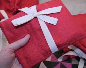 May include: A red fabric gift bag with a white ribbon tied in a bow. The bag is folded and has a herringbone pattern.