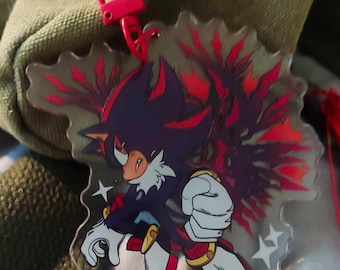 May include: A clear acrylic charm featuring a stylized character with black, red, and white colors. The character is in a dynamic pose with a red and pink clasp attached. The charm has a star design.