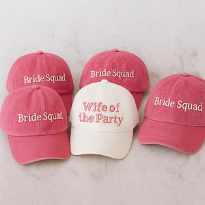 May include: Five baseball caps, four in a vibrant pink and one in white. The pink caps are embroidered with "Bride Squad," while the white cap reads "Wife of the Party." These caps are perfect for a bachelorette party or bridal shower.