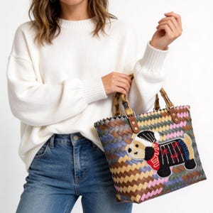 May include: A woven tote bag with a colorful chevron pattern and bamboo handles. The bag features an embroidered dog wearing a hat and scarf. The bag is a mix of brown, blue, green, and pink.
