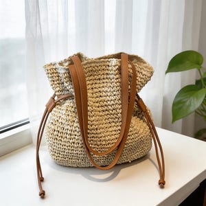 Puede incluir: Bolso tote de paja tejida beige con asas de cuero marr&oacute;n y cierre de cord&oacute;n. El bolso tiene una base redonda. Las asas son lo suficientemente largas para llevarlo al hombro.