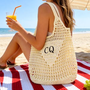 Könnte beinhalten: Eine cremefarbene, offen gewebte Häkeltasche mit den Initialen "CQ" auf der Vorderseite. Die Tasche liegt auf einem rot-weiß gestreiften Strandtuch. Eine Person sitzt am Strand mit einem Getränk und Sandalen.