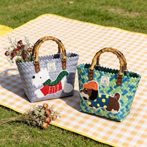 Könnte beinhalten: Zwei gewebte Tragetaschen mit Bambusgriffen. Eine ist grau mit einer weißen Hundeapplikation, die einen rot-grünen Pullover trägt. Die andere ist blau und grün mit einer braunen Hundeapplikation, die einen blauen Polka-Dot-Pullover und einen orangefarbenen Hut trägt. Beide haben ein kleines Etikett.