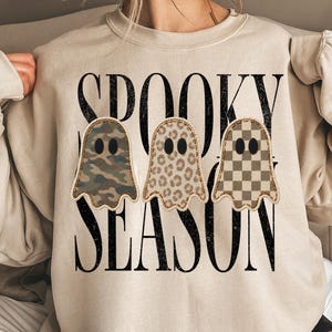 May include: A beige crewneck sweatshirt featuring the black text SPOOKY SEASON and three ghost illustrations. The ghosts have distinct patterns: one with camouflage, one with a leopard print, and one with a brown and beige checkerboard pattern.