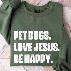 May include: An olive green sweatshirt with the text "PET DOGS. LOVE JESUS. BE HAPPY." in white block letters. The sweatshirt is folded, with the sleeves visible. The fabric appears soft and comfortable.