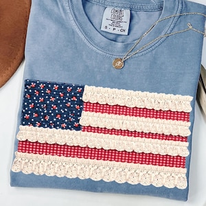 May include: A blue Comfort Colors t-shirt with an American flag design. The flag features a navy blue floral print for the stars and red gingham stripes with cream lace trim. A gold necklace with a pendant rests on the shirt.