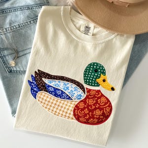 May include: Cream-colored t-shirt featuring an embroidered duck design. The duck has a green polka-dotted head, a yellow beak, and wings with various patterned fabrics in blue, brown, and tan. The shirt is folded on top of blue jeans, with a necklace and a tan hat.