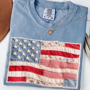 May include: A light blue Comfort Colors t-shirt with a sewn-on American flag design. The flag features white stars on a blue field and red and white stripes made of various patterned fabrics. A gold necklace with a pendant rests on the shirt.