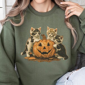 May include: Olive green sweatshirt featuring a Halloween-themed graphic. The graphic depicts three kittens gathered around a carved jack-o'-lantern. The kittens have various markings and colors, and the pumpkin has a smiling face.