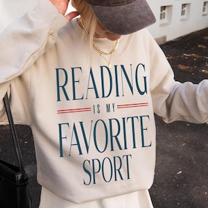 May include: Beige sweatshirt with the text "READING IS MY FAVORITE SPORT" in navy blue. The sweatshirt has a crew neck and long sleeves. A brown baseball cap with white text is worn on the head. A black purse is on the left shoulder.