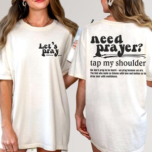 May include: Cream-colored t-shirt with black text. The front reads "Let's Pray" with a Bible verse reference. The back says "Need Prayer? Tap my shoulder" with a supporting quote. A casual, faith-based apparel item.