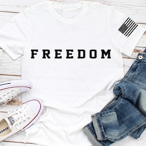 May include: White t-shirt with the word "FREEDOM" in bold black letters. The shirt has a small American flag graphic on the sleeve. The shirt is paired with blue denim jeans and white canvas sneakers.