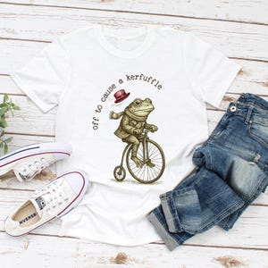 May include: White t-shirt with a frog riding a penny-farthing bicycle. The frog wears a coat, scarf, and hat. The text on the shirt says "off to cause a kerfuffle". A pair of blue jeans and white trainers are also in the image.