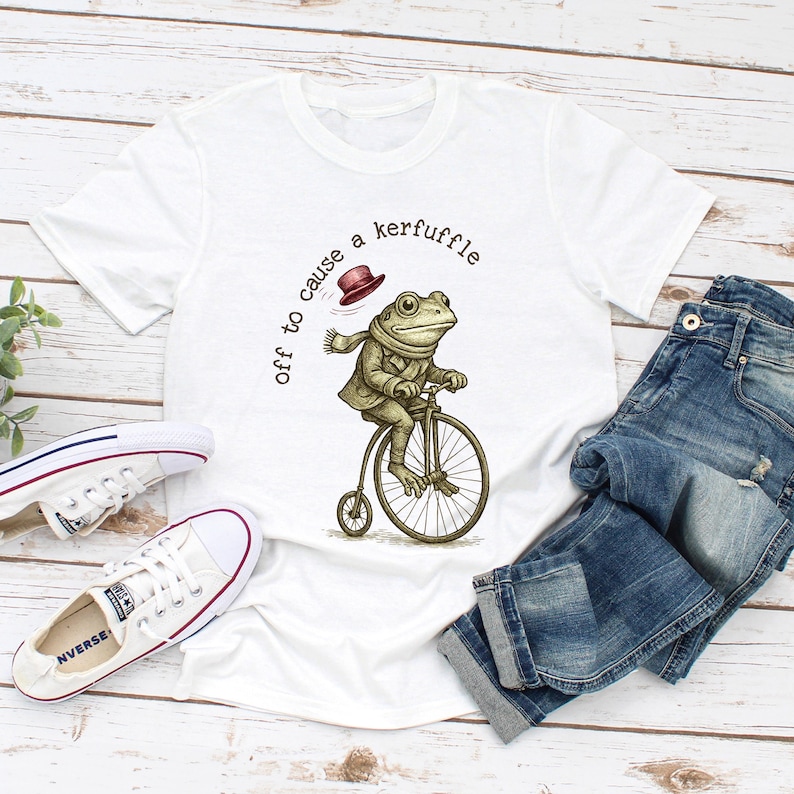 May include: White t-shirt with a frog riding a penny-farthing bicycle. The frog wears a coat, scarf, and hat. The text on the shirt says "off to cause a kerfuffle". A pair of blue jeans and white trainers are also in the image.
