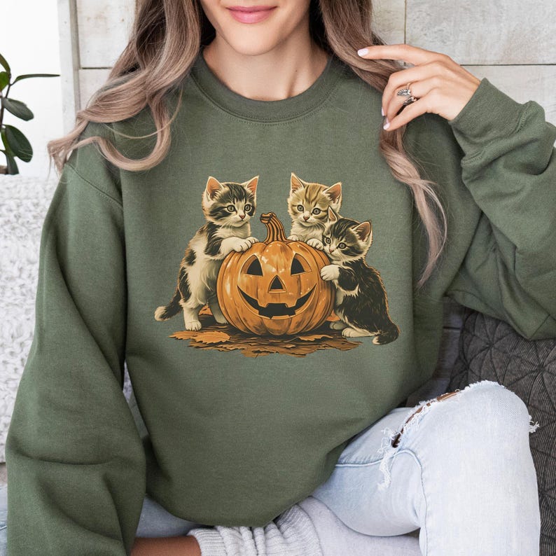 May include: Olive green sweatshirt featuring a Halloween-themed graphic. The graphic depicts three kittens gathered around a carved jack-o'-lantern. The kittens have various markings and colors, and the pumpkin has a smiling face.