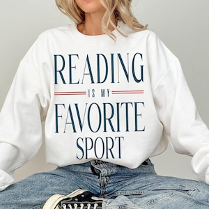 May include: White sweatshirt with the text "READING IS MY FAVORITE SPORT" in navy blue, with red lines separating the phrases. The sweatshirt is a casual style, suitable for everyday wear.