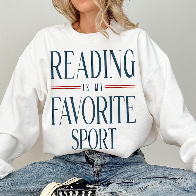 May include: White sweatshirt with the text "READING IS MY FAVORITE SPORT" in navy blue, with red lines separating the phrases. The sweatshirt is a casual style, suitable for everyday wear.