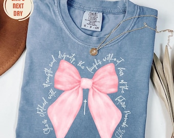 May include: A blue short-sleeve t-shirt featuring a large pink bow graphic in the center. The text surrounding the bow reads, "She is clothed with strength and dignity, she laughs without fear of the future. Proverbs 31:25," with a small cross.