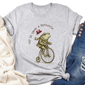 May include: A light grey t-shirt with a quirky illustration of a frog on a penny-farthing bicycle. The frog is dressed in a coat, scarf, and hat. The text above reads "off to cause a kerfuffle."