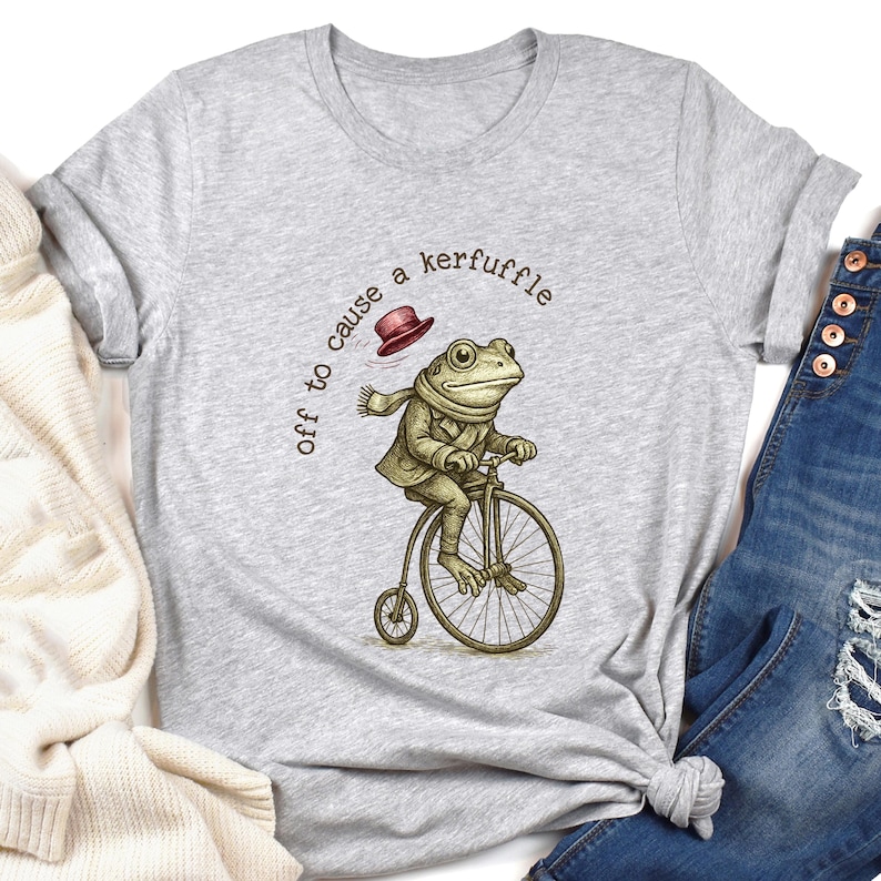 May include: A light grey t-shirt with a quirky illustration of a frog on a penny-farthing bicycle. The frog is dressed in a coat, scarf, and hat. The text above reads "off to cause a kerfuffle."
