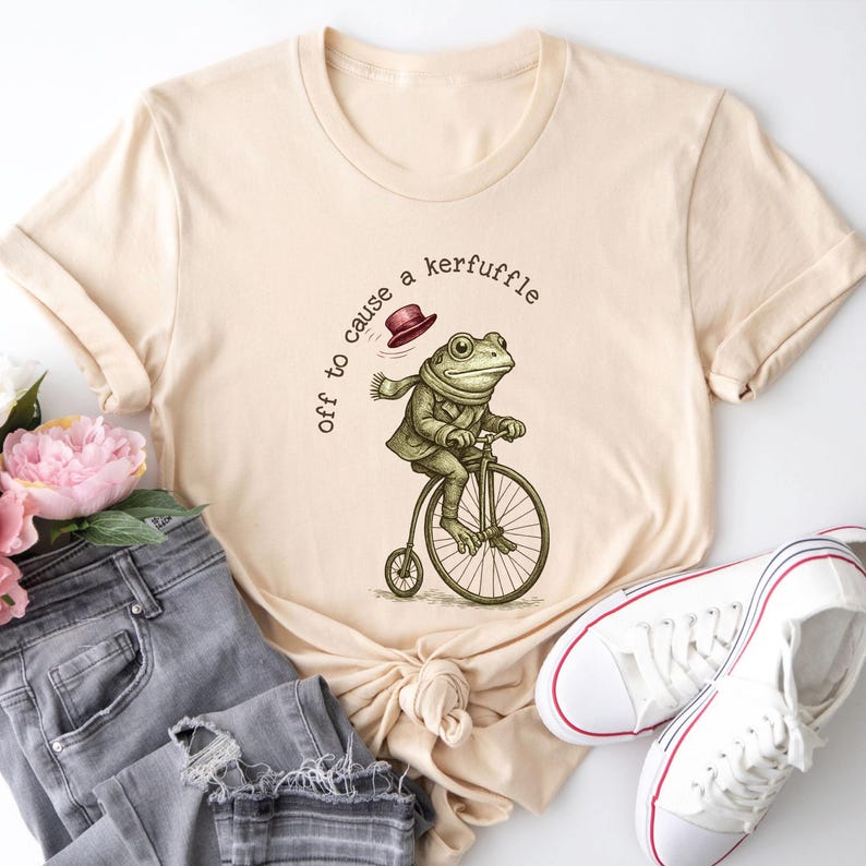 May include: Cream-coloured t-shirt with a quirky frog illustration riding a penny-farthing bicycle. The frog is dressed in a jacket, scarf, and hat. The text above the frog reads "off to cause a kerfuffle."