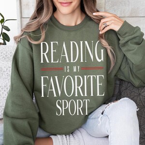 May include: An olive green sweatshirt with the text "READING IS MY FAVORITE SPORT" in white, with two red lines separating the phrases. The sweatshirt is a casual style, suitable for everyday wear.