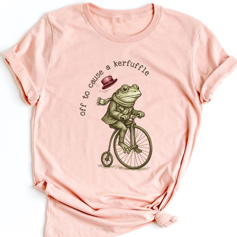 May include: Pale pink t-shirt with a humorous illustration of a frog on a penny-farthing bicycle. The frog is dressed in a coat, scarf, and hat. The text above the frog reads "off to cause a kerfuffle."