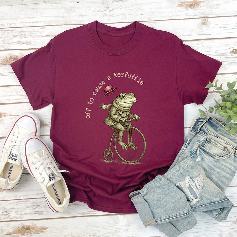 May include: Burgundy t-shirt with a frog riding a penny-farthing bicycle illustration and the text "off to cause a kerfuffle." The image includes light-wash denim jeans and white canvas trainers.