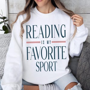 May include: White sweatshirt with the text "READING IS MY FAVORITE SPORT" in navy blue lettering. The text is separated by two red lines. The sweatshirt is a casual style.