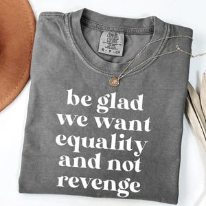 May include: A charcoal gray short-sleeve t-shirt with a crew neckline and white text printed on the front that reads, be glad we want equality and not revenge. The shirt features a Comfort Colors brand label inside the collar indicating size small.