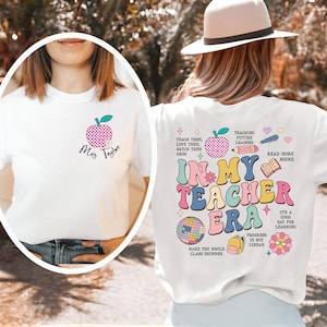 May include: White t-shirts with teacher-themed graphics. One shirt features a pink apple with the name "Mrs. Taylor." The other shirt says "In My Teacher Era" with colorful illustrations of a disco ball, books, and a backpack.
